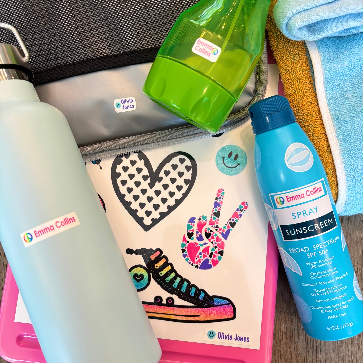 Assorted items with custom name labels including a water bottle, green spray bottle, and blue sunscreen spray on a wooden surface.
