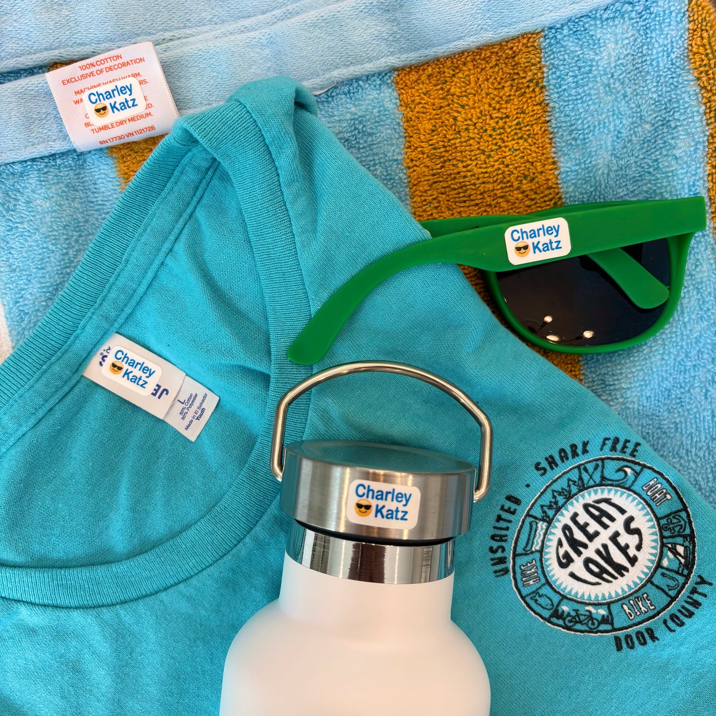 Teal shirt, white water bottle with silver lid, green sunglasses, and towel with 'Charley Katz' branding on a blue towel.