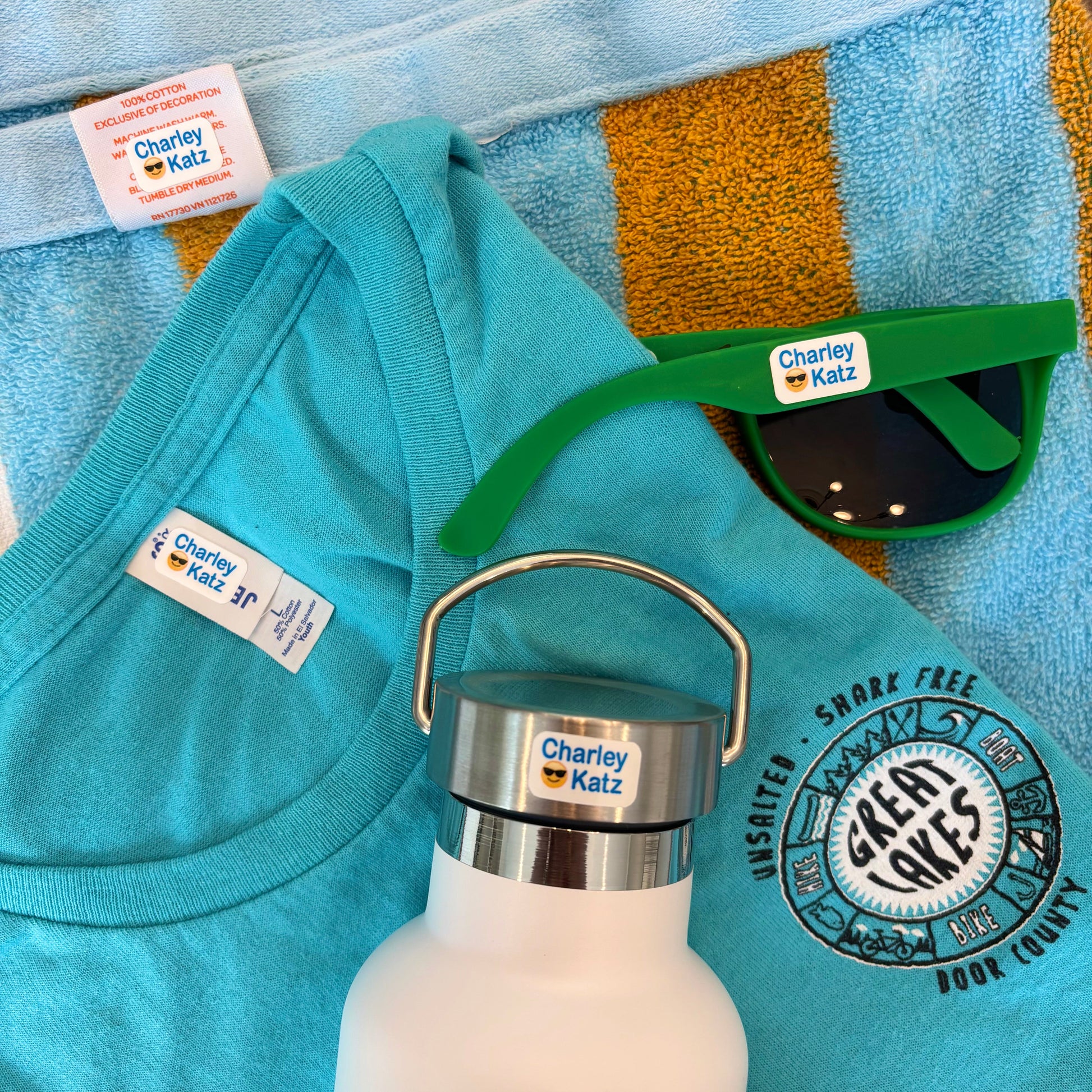 Teal shirt, white water bottle with silver lid, green sunglasses, and towel with 'Charley Katz' branding on a blue towel.
